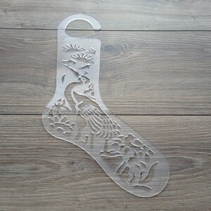 May include: A clear plastic stencil shaped like a stocking with a peacock design. The stencil has a hole at the top for hanging.