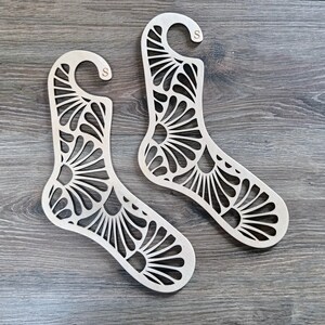 Può includere: Due appendini per calzini in legno con un motivo floreale decorativo. Gli appendini hanno la forma di un calzino e hanno un gancio nella parte superiore.