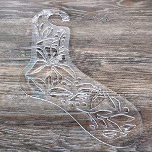 May include: A clear plastic sock-shaped template with a floral design. The template has a small hole at the top for hanging.