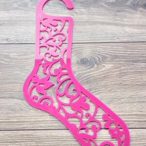 Può includere: Un appendino per calzini in plastica rosa con un design floreale decorativo. L'appendino ha un gancio in alto per appenderlo.