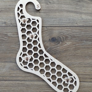 May include: Wooden sock hanger shaped like a sock with a honeycomb pattern and four small bee images. The hanger has a hook at the top and the letter "S" is engraved on the top of the hook.