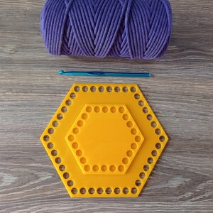 Op de afbeelding: Twee gele, zeshoekige plastic haaksteekmarkeerders, een binnen de andere, met een blauwe haaknaald en een bolletje paars garen op een houten oppervlak.