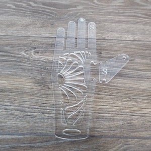 Könnte beinhalten: Eine transparente Hand-Schablone aus Kunststoff mit einem dekorativen Design und dem Buchstaben "S" an den Fingern und dem Daumen. Die Schablone ist zum Basteln und Dekorieren gedacht.