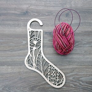 Può includere: Un porta-ferri da maglia in legno a forma di calza con un motivo floreale. Il porta-ferri ha un gancio in alto e la lettera "S" è incisa sul lato. Sullo sfondo c'è un gomitolo di filo rosa con due ferri da maglia.