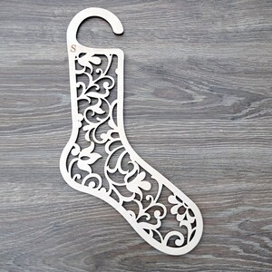 Può includere: Un appendi calze in legno bianco con un design floreale decorativo. L'appendi calze ha un gancio in alto e la lettera "S" è incisa sul lato.