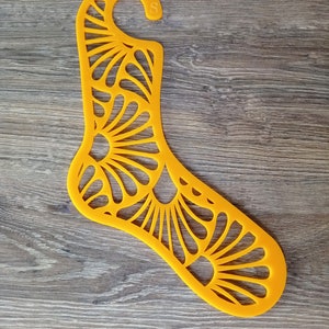 May include: A yellow plastic sock hanger with a decorative floral design. The hanger has a hook at the top and the letter "S" printed on the side.