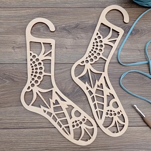 Può includere: Due appendini per calzini in legno con un motivo floreale decorativo. Gli appendini hanno la forma di calzini e hanno un foro nella parte superiore per appenderli.