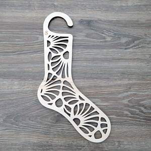 Può includere: Un appendino per calzini in legno con un design decorativo a taglio. L'appendino è etichettato con la lettera "S" ed è progettato per contenere calzini.