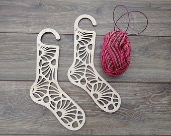 Könnte beinhalten: Zwei hölzerne Socken-förmige Stricknadelhalter mit einem dekorativen Blumenmuster. Die Halter sind mit "S" gekennzeichnet und werden auf einer Holzoberfläche mit einer Knäuel rosa Garn gezeigt.