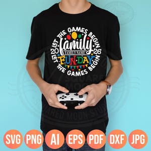 May include: Black t-shirt with a colorful graphic that reads "Let the Games Begin" with "Family Fun Day" in the center. The design includes balloons, pennants, and the year "2026". A person is holding a white game controller.