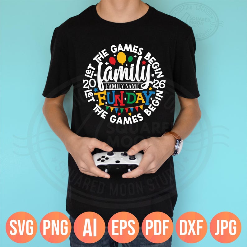 May include: Black t-shirt with a colorful graphic that reads "Let the Games Begin" with "Family Fun Day" in the center. The design includes balloons, pennants, and the year "2026". A person is holding a white game controller.