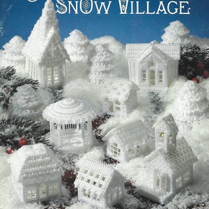 Może przedstawiać: Broszura ze wzorami zatytułowana Plastic Canvas Snow Village od American School of Needlework, przedstawiająca białe konstrukcje z plastikowej kanwy, w tym domy, kościół i altanę, umieszczone na sztucznym śniegu.