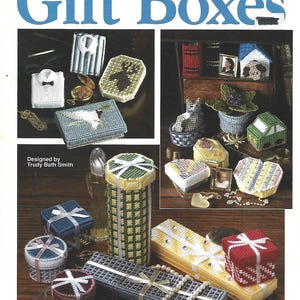 Peut inclure: Un livret de modèles artisanaux intitulé Plastic Canvas Gift Boxes par The Needlecraft Shop. La couverture présente diverses petites boîtes et contenants décoratifs cousus en canevas plastique de formes, couleurs et motifs variés.