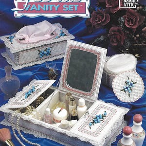 May include: A plastic canvas vanity set featuring a tissue box cover, a lidded mirror box, and a round container, all decorated with white mesh, pink trim, and blue beaded floral accents. The set includes text reading Beaded Floral Vanity Set.