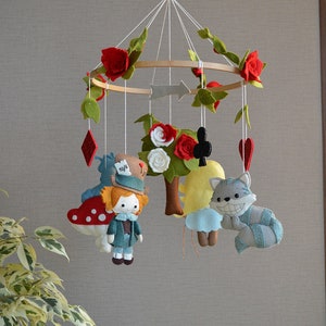 May include: A felt baby mobile with a whimsical Alice in Wonderland theme. It features characters like the Mad Hatter, the Cheshire Cat, and Alice herself, along with red roses, playing cards, and a tree.