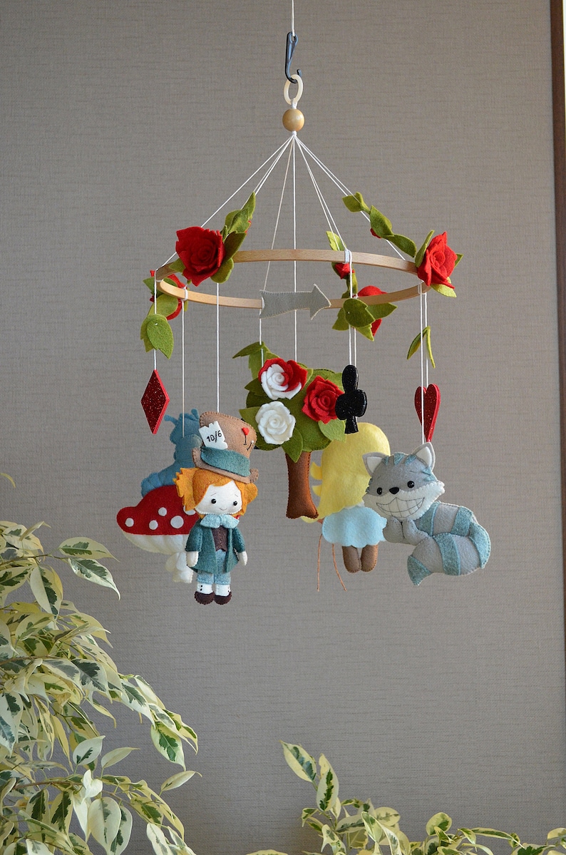 May include: A felt baby mobile with a whimsical Alice in Wonderland theme. It features characters like the Mad Hatter, the Cheshire Cat, and Alice herself, along with red roses, playing cards, and a tree.