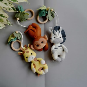 May include: A baby mobile with felt safari animals, including a giraffe, monkey, and zebra. The animals are suspended from wooden rings with felt leaves. The giraffe is yellow with brown spots, the monkey is brown, and the zebra is white with grey stripes.