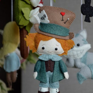 May include: A felt toy of the Mad Hatter from Alice in Wonderland. He is wearing a brown and green hat with the number 10/6 on it. He has orange hair and a white face with black eyes. He is wearing a green jacket and blue pants.