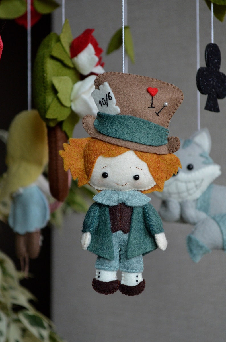 May include: A felt toy of the Mad Hatter from Alice in Wonderland. He is wearing a brown and green hat with the number 10/6 on it. He has orange hair and a white face with black eyes. He is wearing a green jacket and blue pants.
