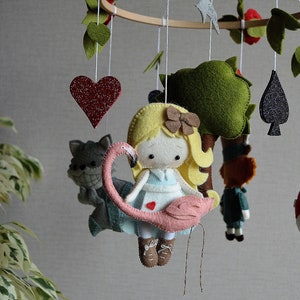 May include: A felt baby mobile with a pink flamingo, a girl with blonde hair, a green tree, a red heart, a black spade, and a gray cat. The mobile is hanging from a wooden ring.