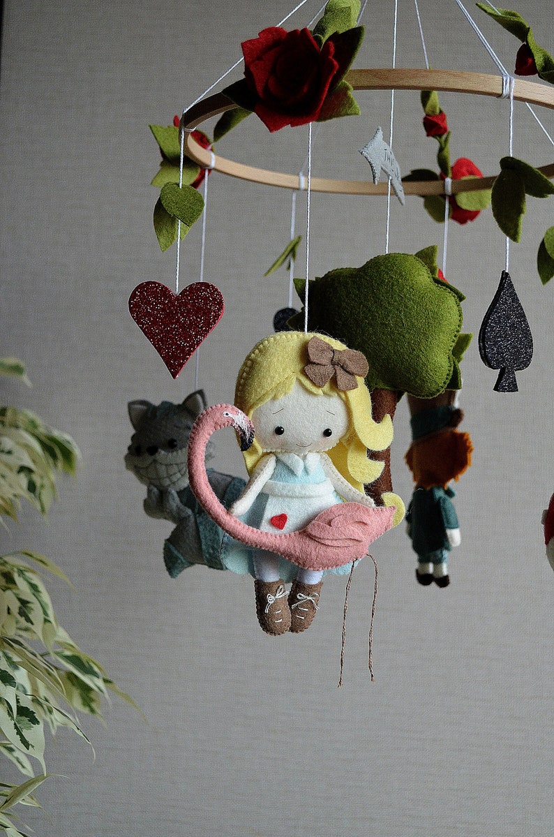 May include: A felt baby mobile with a pink flamingo, a girl with blonde hair, a green tree, a red heart, a black spade, and a gray cat. The mobile is hanging from a wooden ring.