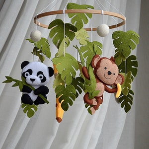 May include: A felt baby mobile with a panda, monkey, and banana hanging from a wooden ring. The mobile is decorated with green felt leaves.