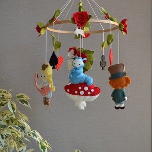 May include: A wooden baby mobile with felt characters from Alice in Wonderland. The mobile features Alice, the Mad Hatter, the Cheshire Cat, and playing cards. The mobile is decorated with red roses and green leaves.