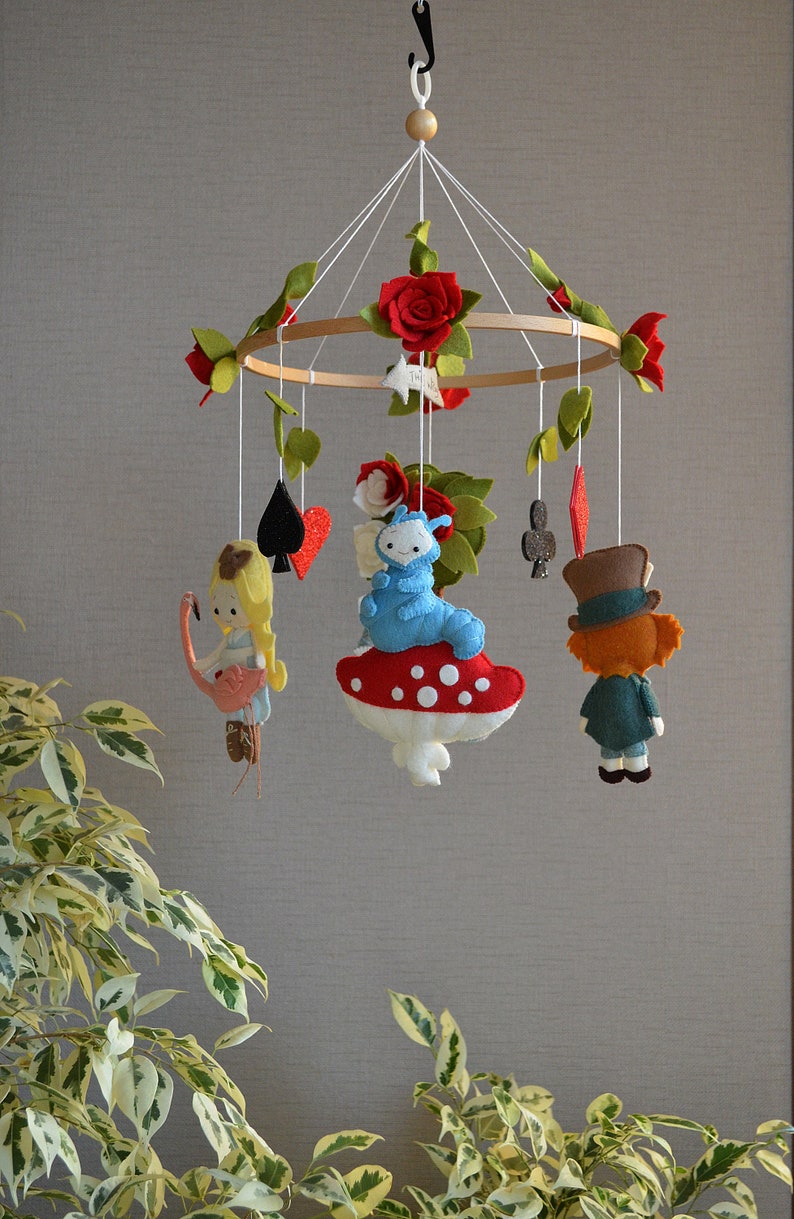 May include: A wooden baby mobile with felt characters from Alice in Wonderland. The mobile features Alice, the Mad Hatter, the Cheshire Cat, and playing cards. The mobile is decorated with red roses and green leaves.