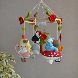 May include: A felt baby mobile with a whimsical Alice in Wonderland theme. It features a red and white mushroom, a blue caterpillar, a playing card, a grey rabbit, a red heart, a red rose, and a felt doll of Alice riding a pink flamingo. The mobile is decorated with green leaves and red roses.