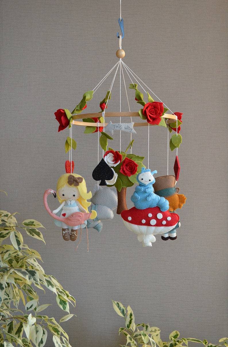 May include: A felt baby mobile with a whimsical Alice in Wonderland theme. It features a red and white mushroom, a blue caterpillar, a playing card, a grey rabbit, a red heart, a red rose, and a felt doll of Alice riding a pink flamingo. The mobile is decorated with green leaves and red roses.