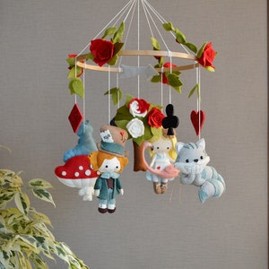 May include: A felt baby mobile with a whimsical Alice in Wonderland theme. It features characters like the Mad Hatter, Alice, the Cheshire Cat, and a playing card. The mobile is decorated with red roses and green leaves.