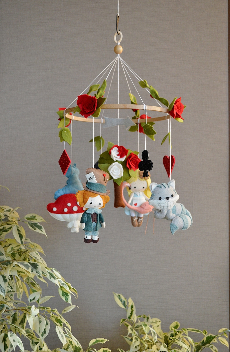 May include: A felt baby mobile with a whimsical Alice in Wonderland theme. It features characters like the Mad Hatter, Alice, the Cheshire Cat, and a playing card. The mobile is decorated with red roses and green leaves.