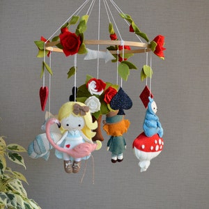 May include: A felt baby mobile with a white, pink, and blue Alice in Wonderland theme. The mobile features Alice holding a pink flamingo, the Mad Hatter, the Queen of Hearts, and the Cheshire Cat.
