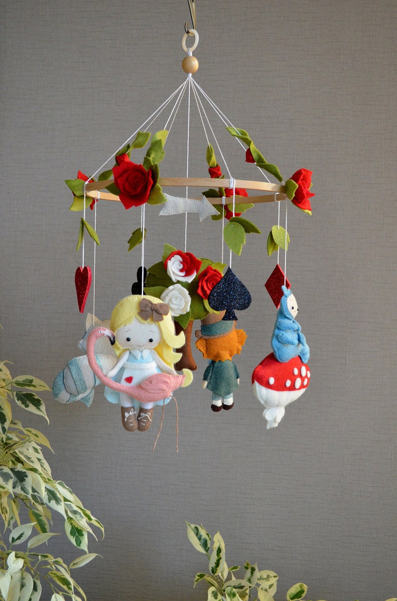 May include: A felt baby mobile with a white, pink, and blue Alice in Wonderland theme. The mobile features Alice holding a pink flamingo, the Mad Hatter, the Queen of Hearts, and the Cheshire Cat.