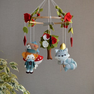 May include: A felt baby mobile with a whimsical Alice in Wonderland theme. It features a red-haired girl with a top hat, a Cheshire cat, a tree with white flowers, and red roses.