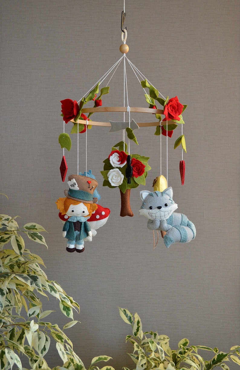 May include: A felt baby mobile with a whimsical Alice in Wonderland theme. It features a red-haired girl with a top hat, a Cheshire cat, a tree with white flowers, and red roses.