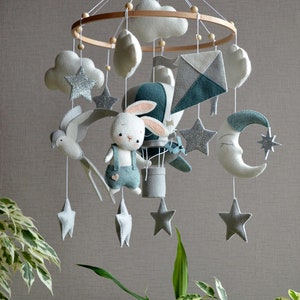 Puede incluir: Un móvil para bebé con un conejito blanco, un globo aerostático, nubes, estrellas y una luna creciente. El móvil está hecho de fieltro y tiene un anillo de madera.