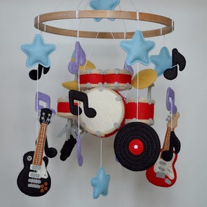 Pode incluir: Um móbile suspenso com instrumentos musicais e símbolos de feltro pendurados numa estrutura circular de madeira. O móbile inclui uma bateria vermelha e branca, uma guitarra elétrica preta, uma guitarra elétrica vermelha, um disco de vinil preto e estrelas azul-claras.