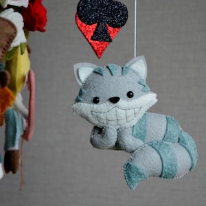 May include: A gray and blue felt cat with a wide grin and a long tail hangs from a mobile. The cat is a whimsical character with a playful expression. The mobile also features a red and black felt playing card with a club symbol.