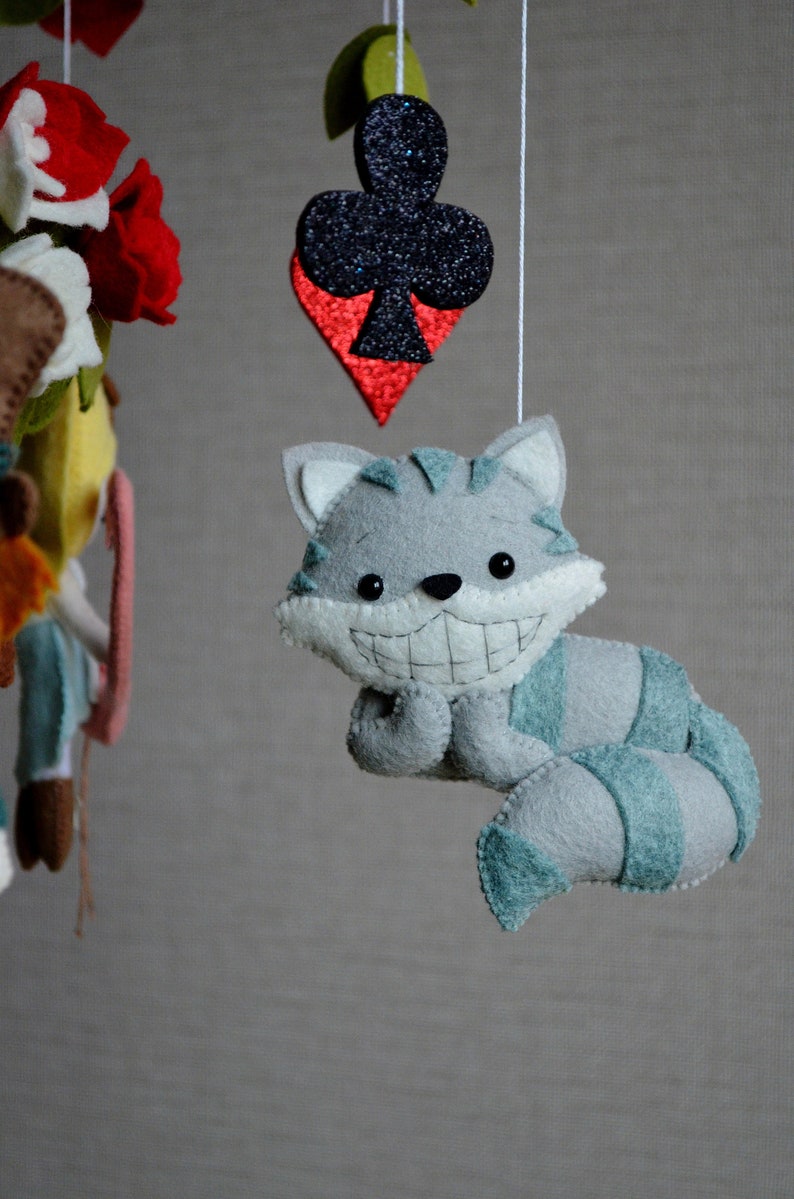 May include: A gray and blue felt cat with a wide grin and a long tail hangs from a mobile. The cat is a whimsical character with a playful expression. The mobile also features a red and black felt playing card with a club symbol.