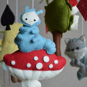 May include: A blue felt caterpillar sits on a red and white mushroom with white polka dots. The mushroom is hanging from a wooden mobile with other felt toys.