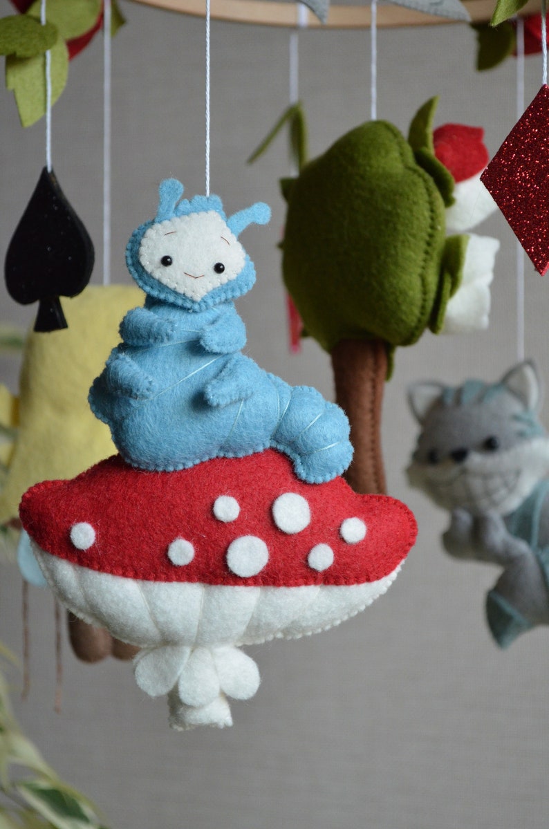 May include: A blue felt caterpillar sits on a red and white mushroom with white polka dots. The mushroom is hanging from a wooden mobile with other felt toys.