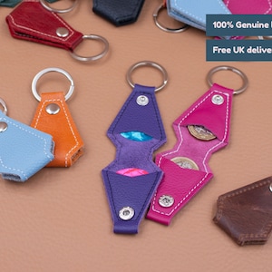 May include: A collection of small, diamond-shaped leather keychains in various colors including light blue, orange, purple, pink, and brown. Each keychain features a metal split ring, a silver snap button, and a small internal pocket for coins.