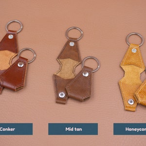 May include: Three sets of leather keychains in conker, mid tan, and honeycomb colors. Each set includes a long, notched leather strap with a metal ring and snap button, paired with a smaller, matching leather pouch keychain with a metal ring.