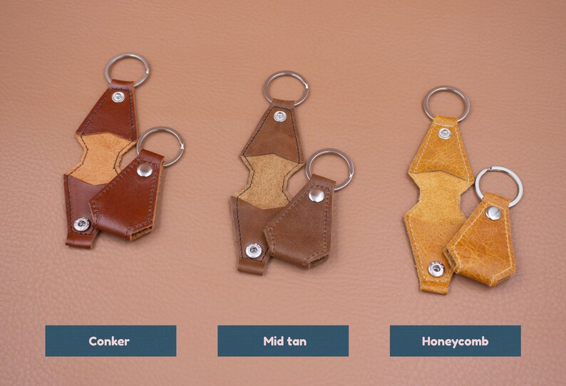 May include: Three sets of leather keychains in conker, mid tan, and honeycomb colors. Each set includes a long, notched leather strap with a metal ring and snap button, paired with a smaller, matching leather pouch keychain with a metal ring.