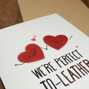May include: A white card with two red felt hearts with smiling faces. The hearts are holding hands and the card says "We're perfect to-leather".