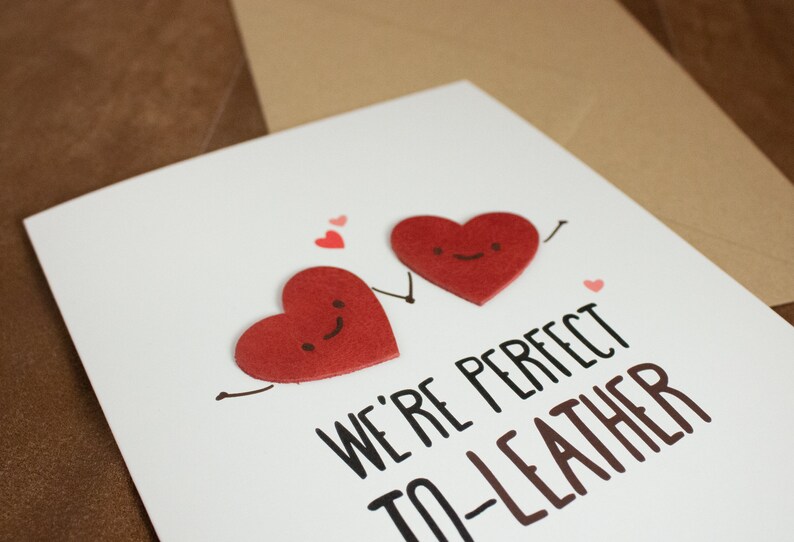 May include: A white card with two red felt hearts with smiling faces. The hearts are holding hands and the card says "We're perfect to-leather".