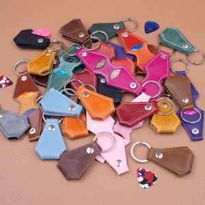 May include: A collection of multi-colored leather guitar pick holders featuring metal snap closures and attached silver-toned key rings. The assortment includes various shades such as pink, blue, brown, green, orange, and black, with several guitar picks.
