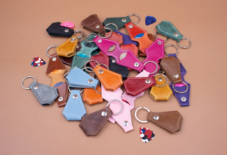 May include: A collection of multi-colored leather guitar pick holders featuring metal snap closures and attached silver-toned key rings. The assortment includes various shades such as pink, blue, brown, green, orange, and black, with several guitar picks.