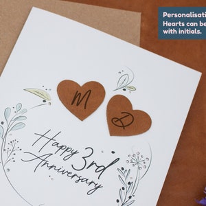 K&ouml;nnte beinhalten: Eine wei&szlig;e Karte mit einem floralen Design und dem Text "Happy 3rd Anniversary". Zwei braune herzf&ouml;rmige Papierschnitte mit den Initialen "M" und "D" sind an der Karte befestigt.